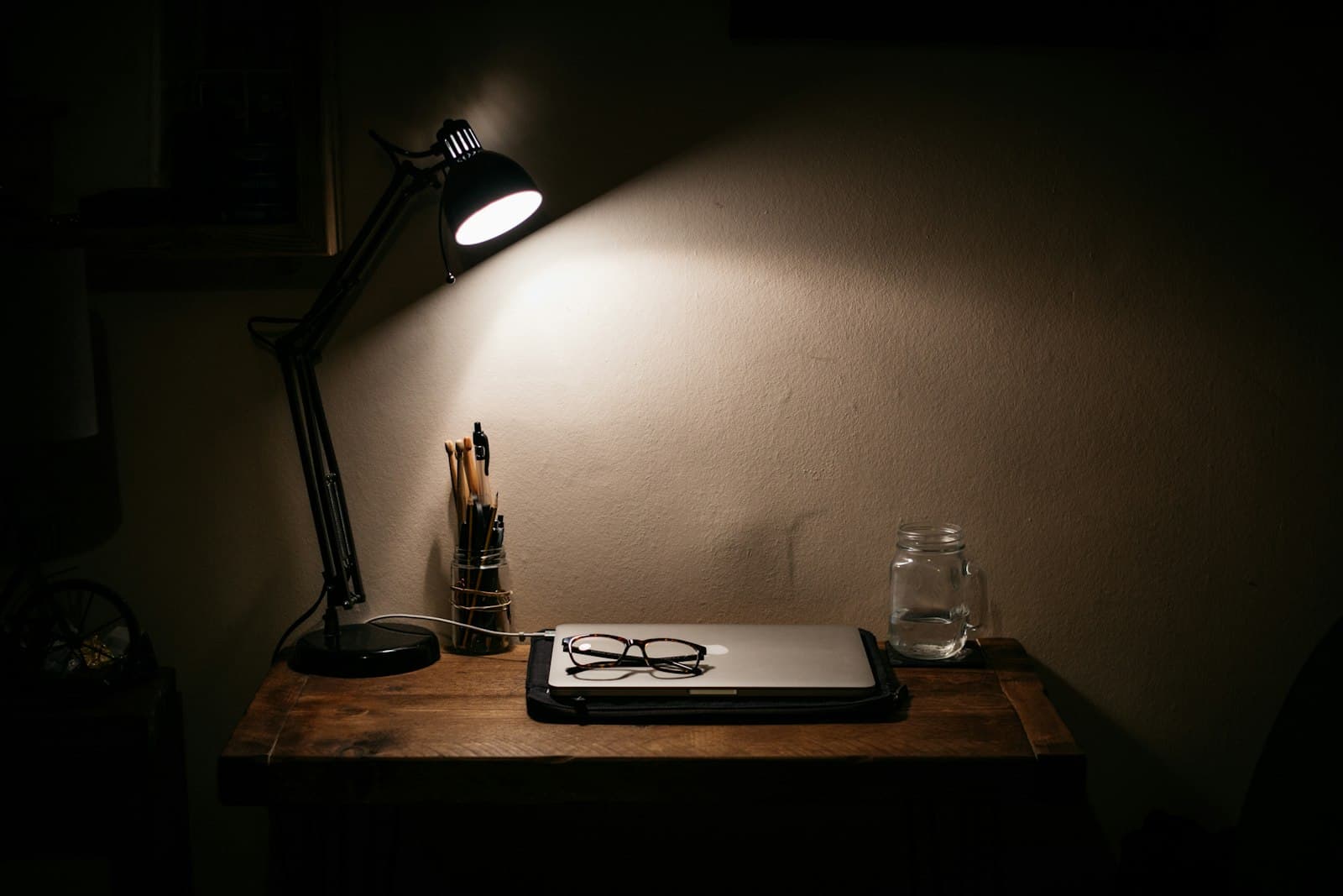 Desk lamp lighting a closed laptop and a jar of pens on a wooden desk